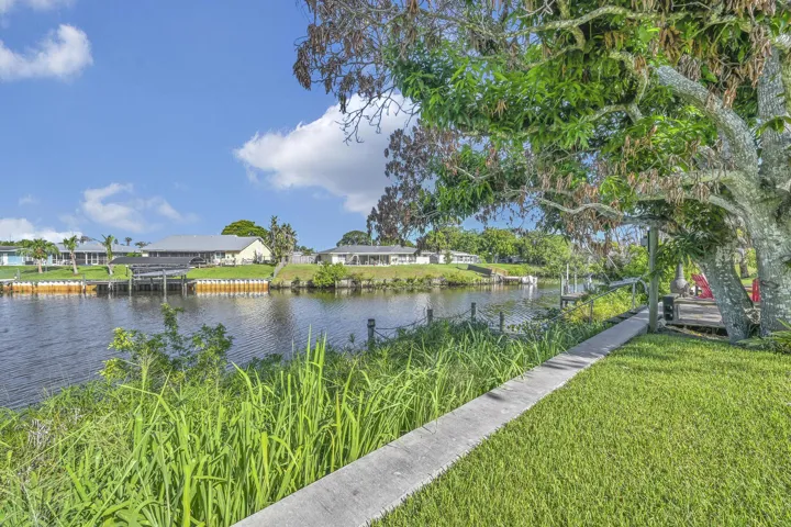 Waterfront canal property featuring a concrete walkway, mature trees, and a wooden dock structure