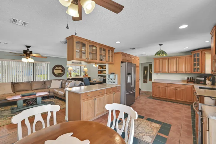 Open-concept kitchen and living area featuring warm wood cabinetry, a central island with countertop seating, stainless steel appliances, and terracotta-style floor tiling with decorative inlays