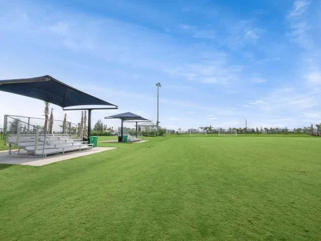 Expansive green field with two shaded spectator seating areas, featuring aluminum bleachers and protective fencing