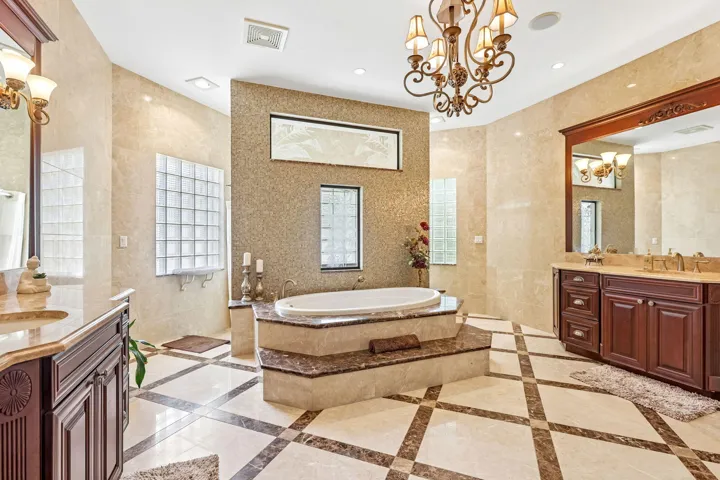 Spacious bathroom featuring a built-in soaking tub with tiered stone surround, decorative mosaic tile wall, two separate vanity cabinets with integrated sinks, and a large central chandelier
