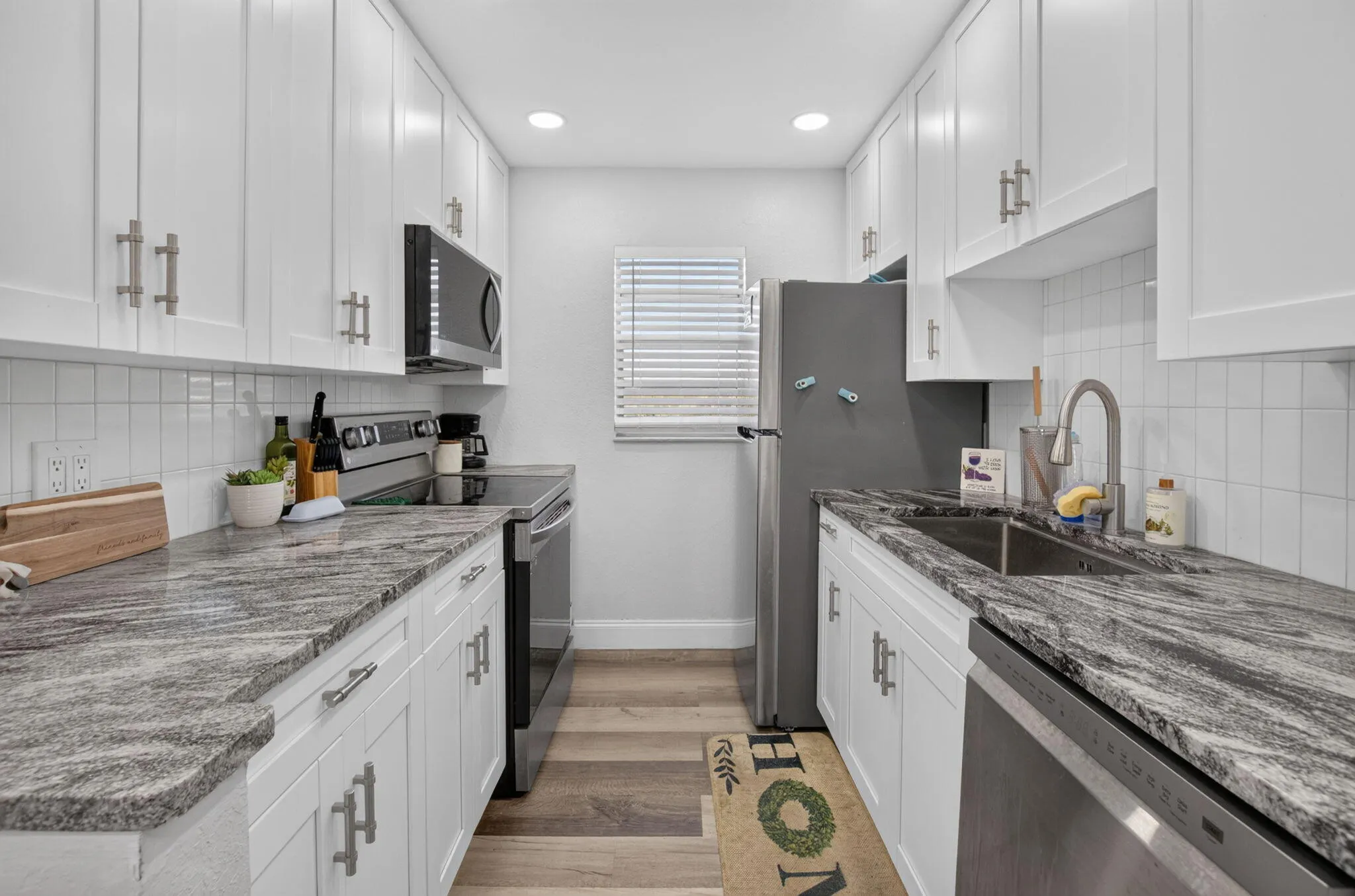 Galley kitchen featuring white shaker cabinetry, stainless steel appliances, patterned countertops, a white tile backsplash, and wood-finish flooring