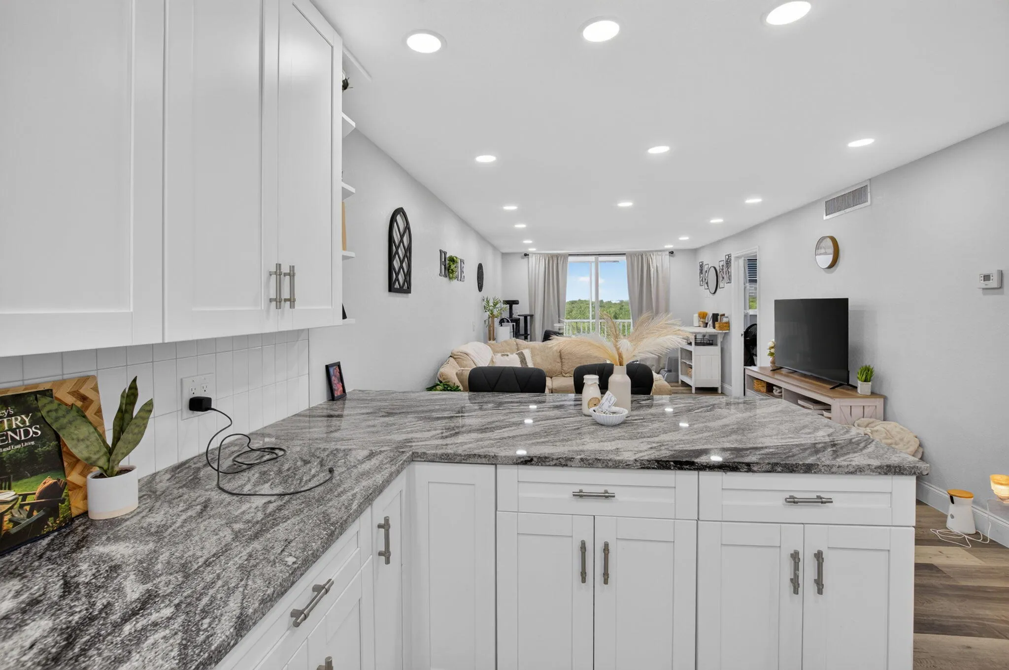 Modern kitchen featuring white shaker cabinetry, patterned stone countertops, white subway tile backsplash, recessed lighting, and wood-finish flooring extending into the living area