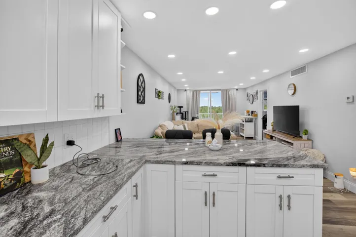 Modern kitchen featuring white shaker cabinetry, patterned stone countertops, white subway tile backsplash, recessed lighting, and wood-finish flooring extending into the living area
