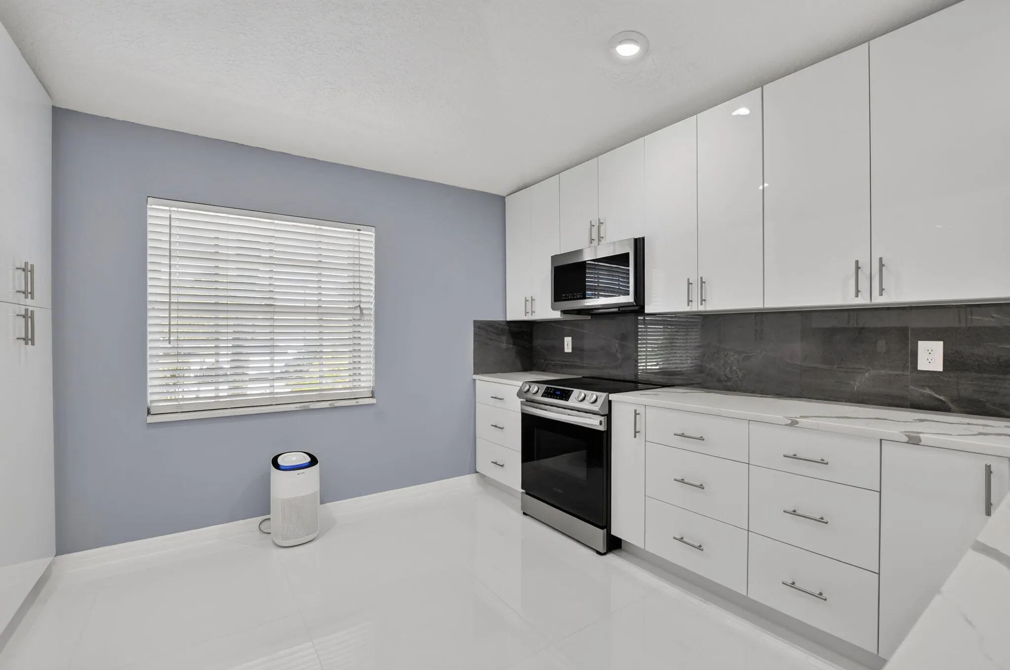 Modern kitchen featuring sleek white cabinetry, stainless steel appliances, a dark stone backsplash, light countertops, and white tile flooring