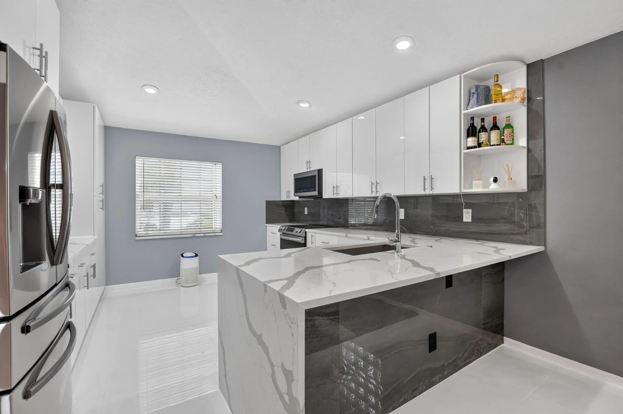 Modern kitchen featuring a waterfall island with a stainless steel sink, sleek white cabinetry, stainless steel appliances, dark stone-look backsplash, and recessed lighting