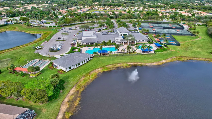 Expansive community amenities including a large clubhouse, multiple swimming pools, extensive tennis courts, bocce ball courts, and a parking area, all bordered by lush landscaping and a serene lake with a fountain