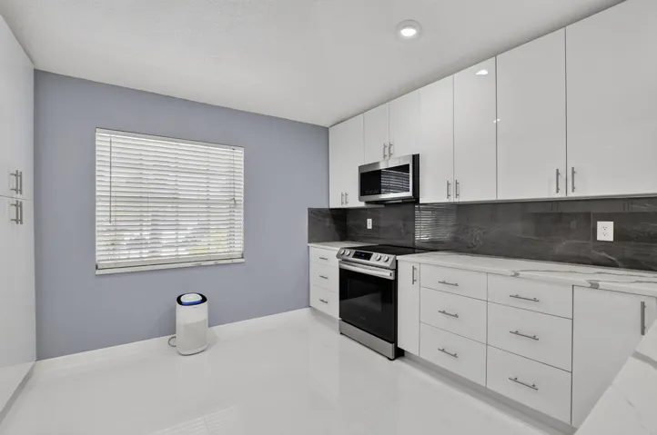 Modern kitchen featuring sleek white cabinetry, stainless steel appliances, a dark stone backsplash, light countertops, and white tile flooring