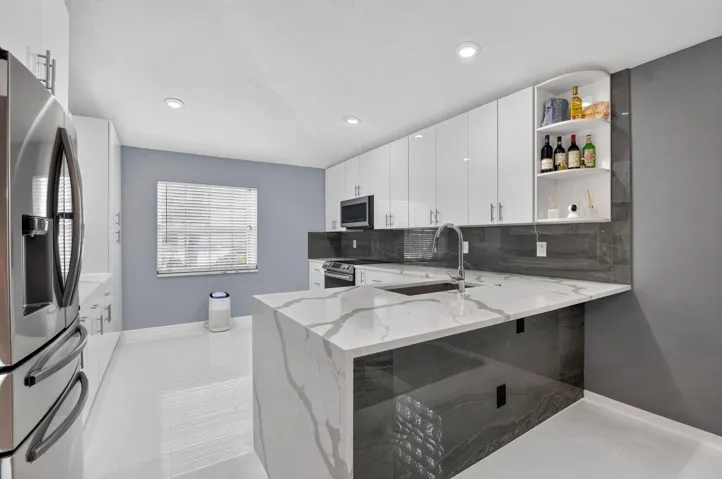 Modern kitchen featuring a waterfall island with a stainless steel sink, sleek white cabinetry, stainless steel appliances, dark stone-look backsplash, and recessed lighting