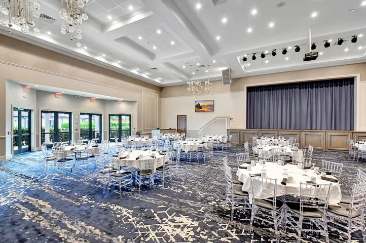 Spacious ballroom featuring a large stage with dark blue curtains, multiple sets of double glass doors, recessed lighting, decorative ceiling fixtures, and a distinct patterned carpet