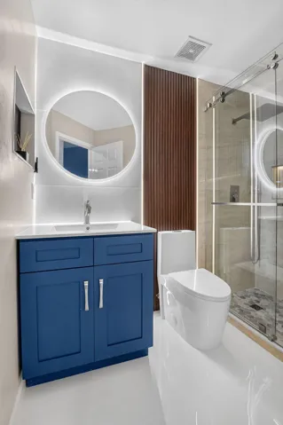 Contemporary bathroom featuring a blue vanity with a white countertop and undermount sink, a backlit circular mirror, a modern toilet, a glass-enclosed shower with mosaic floor tiling, and a vertical wood-finish accent panel