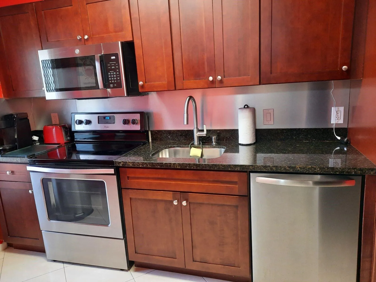 Kitchen featuring wood-finish cabinetry, dark granite-style countertops, stainless steel appliances, stainless steel backsplash, and an undermount sink with a gooseneck faucet