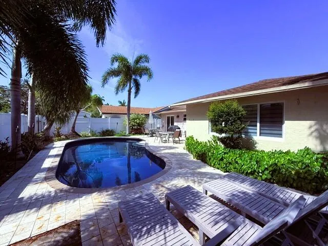 Curved inground pool with stone paver surround, bordered by tropical landscaping, a stucco exterior, and a privacy fence