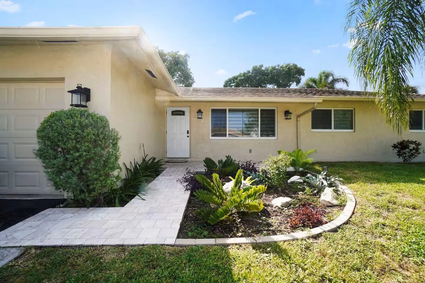 Stucco exterior featuring a tiled walkway, landscaped garden bed, white entry door with frosted glass insert, and a garage with overhead door