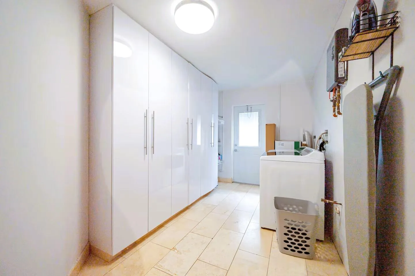 Bright utility space featuring floor-to-ceiling white cabinetry with brushed metal hardware, neutral-toned floor tile, and a rear exit door