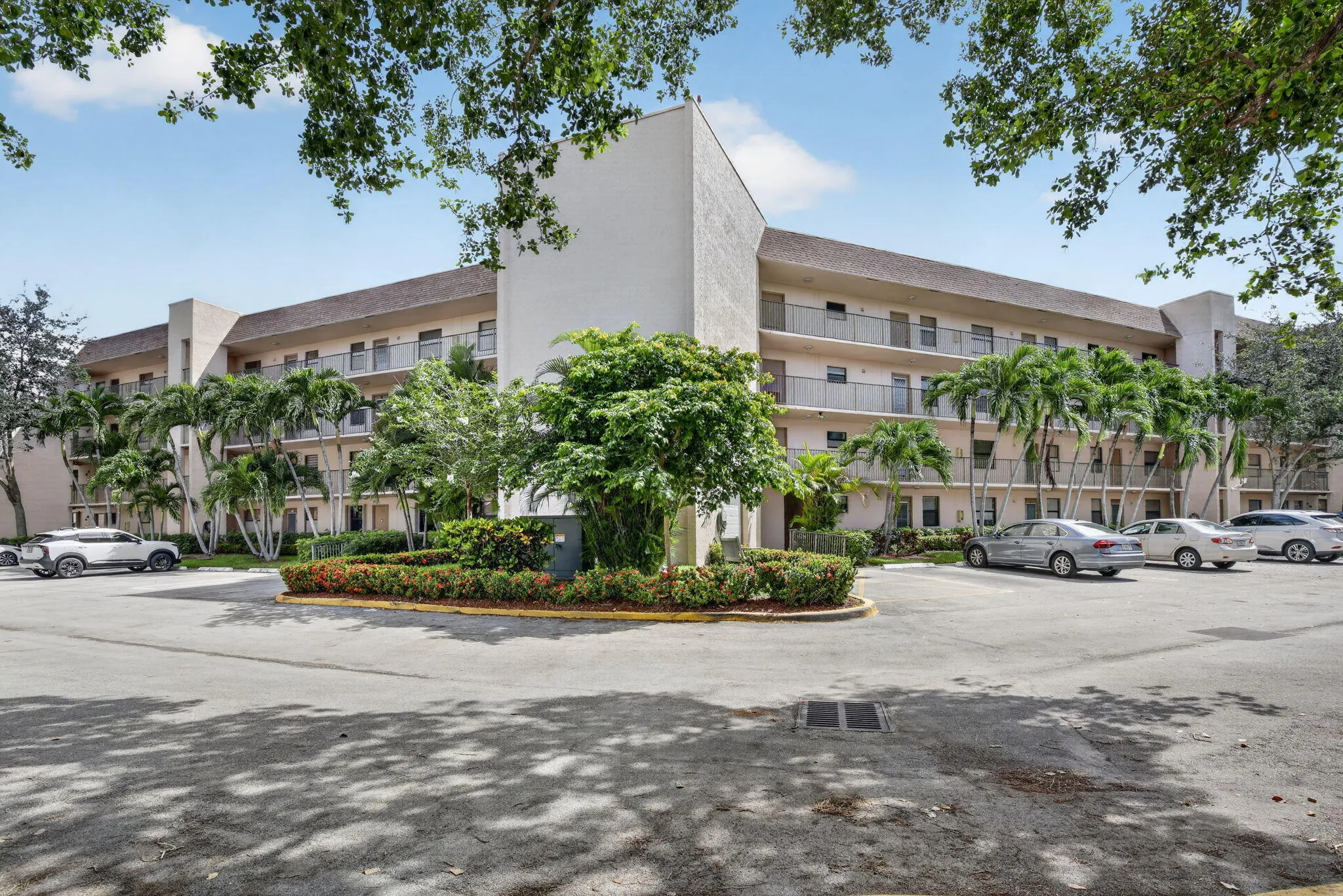 Multi-story building featuring light-colored stucco exterior, balconies with metal railings, and a surrounding paved parking area