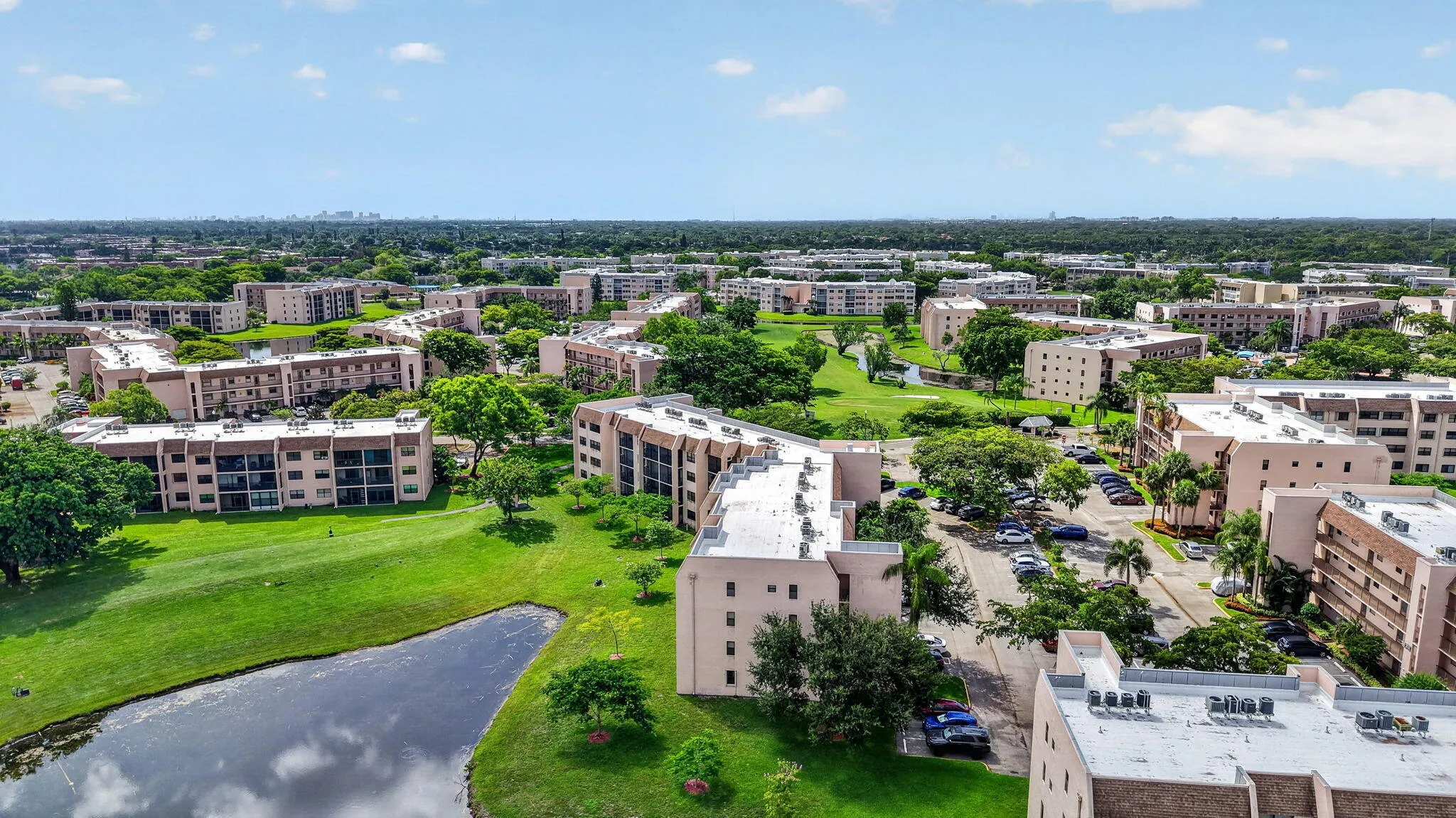 Aerial perspective showcasing multiple residential buildings, lush green spaces, a reflective lake, and paved parking areas