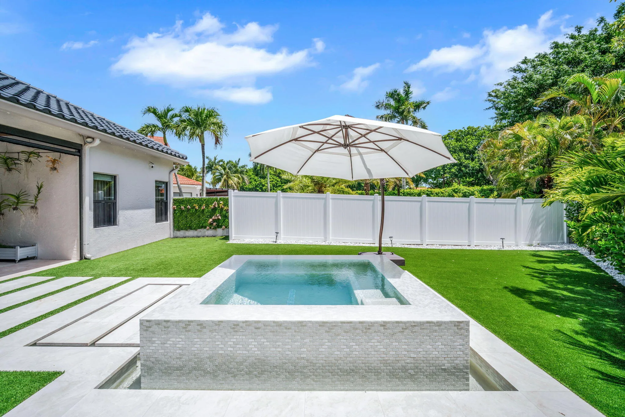 Backyard featuring a tiled spa with integrated steps, artificial turf, white fencing, and concrete paver pathways