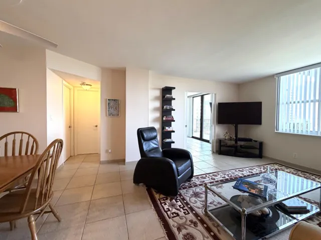 Spacious living area featuring light-toned tile flooring, large windows with vertical blinds, and a built-in shelving unit