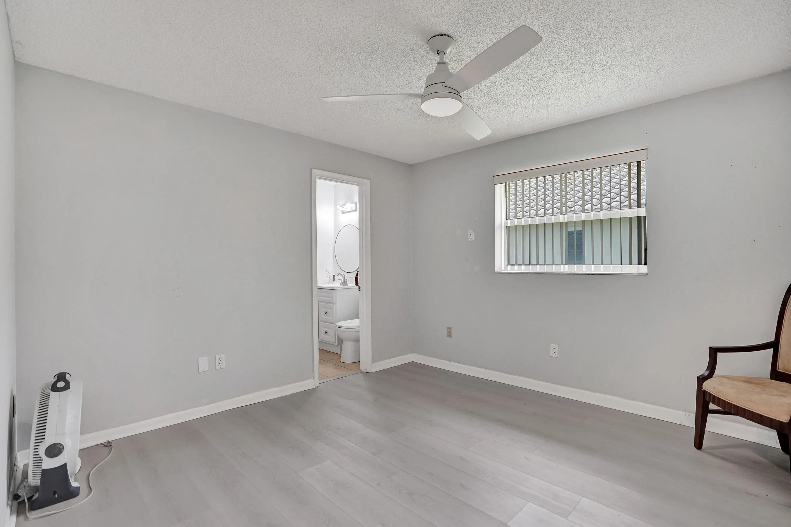 Spacious room featuring light wood-finish flooring, a ceiling fan with integrated lighting, a window with horizontal blinds, and a doorway opening to a bathroom with a white vanity and round mirror