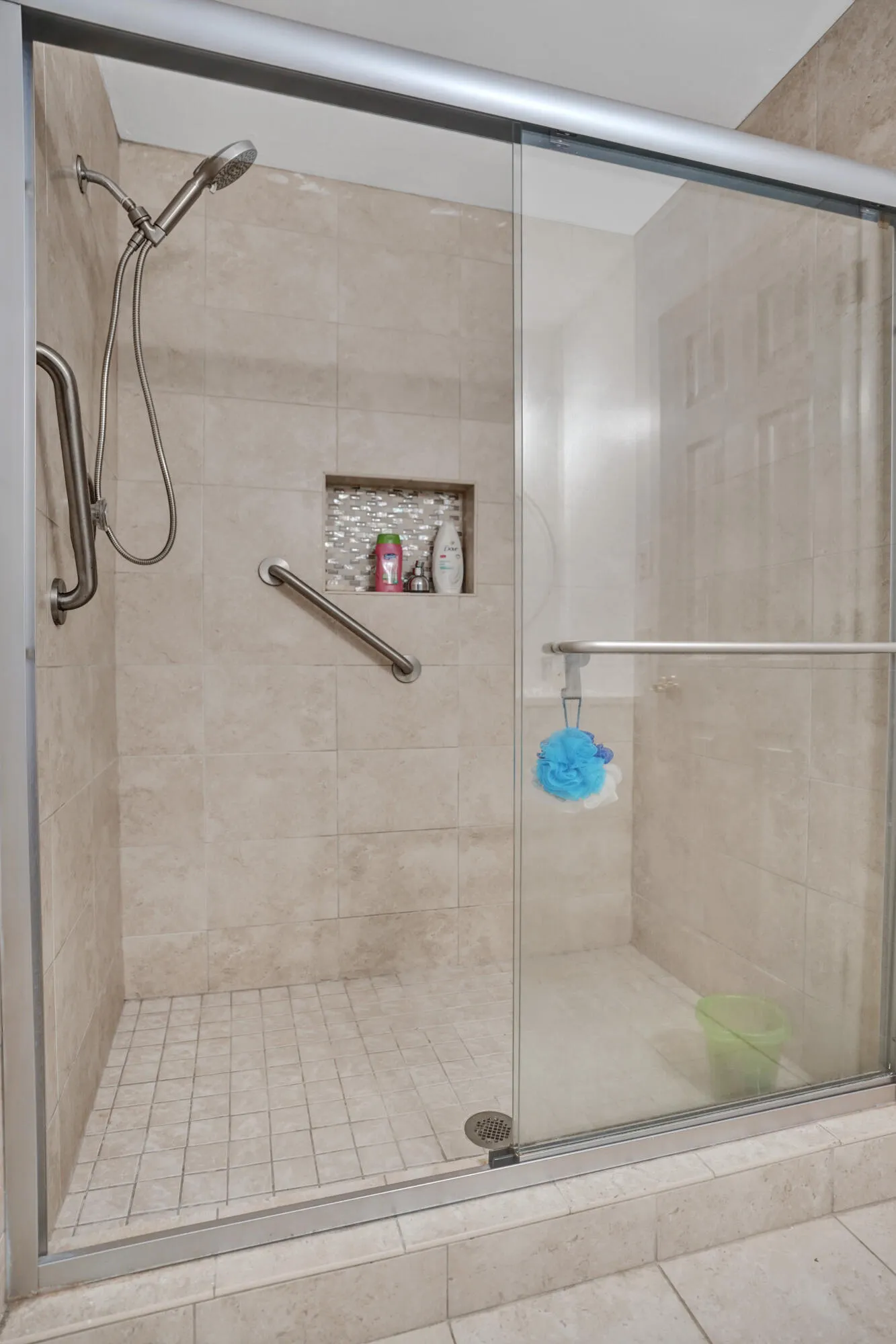Walk-in shower featuring light-toned tile walls, a mosaic-tiled recessed niche, a handheld shower fixture, and multiple grab bars