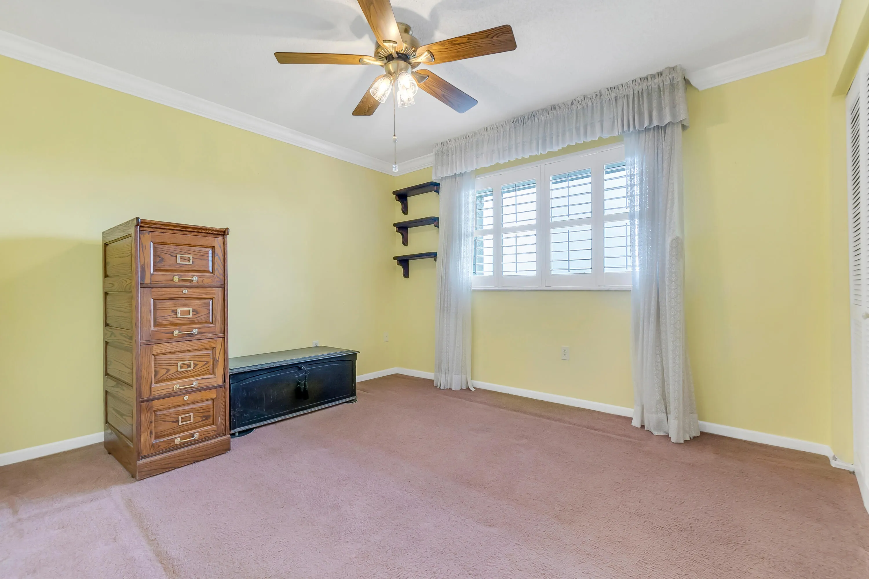 Carpeted room with a ceiling fan light fixture, crown molding, and a window featuring white plantation shutters and sheer lace curtains