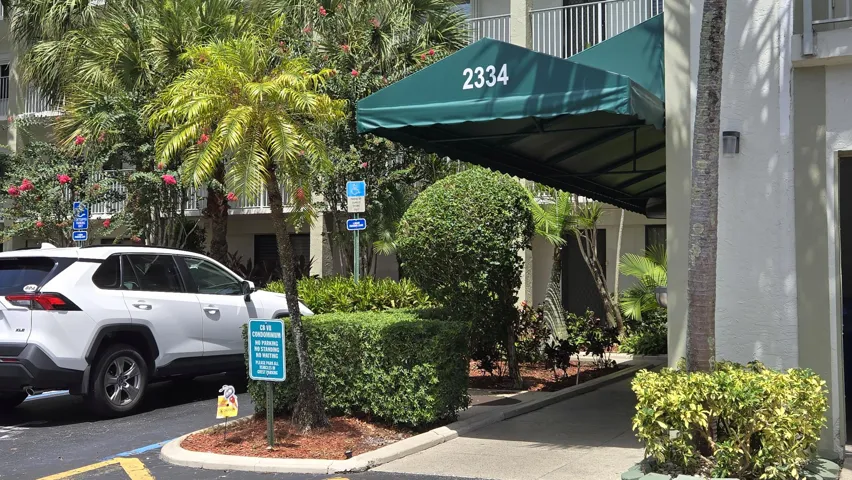 Building entrance featuring a green awning, surrounding lush tropical landscaping with palm trees and manicured hedges, and a dedicated drop-off area