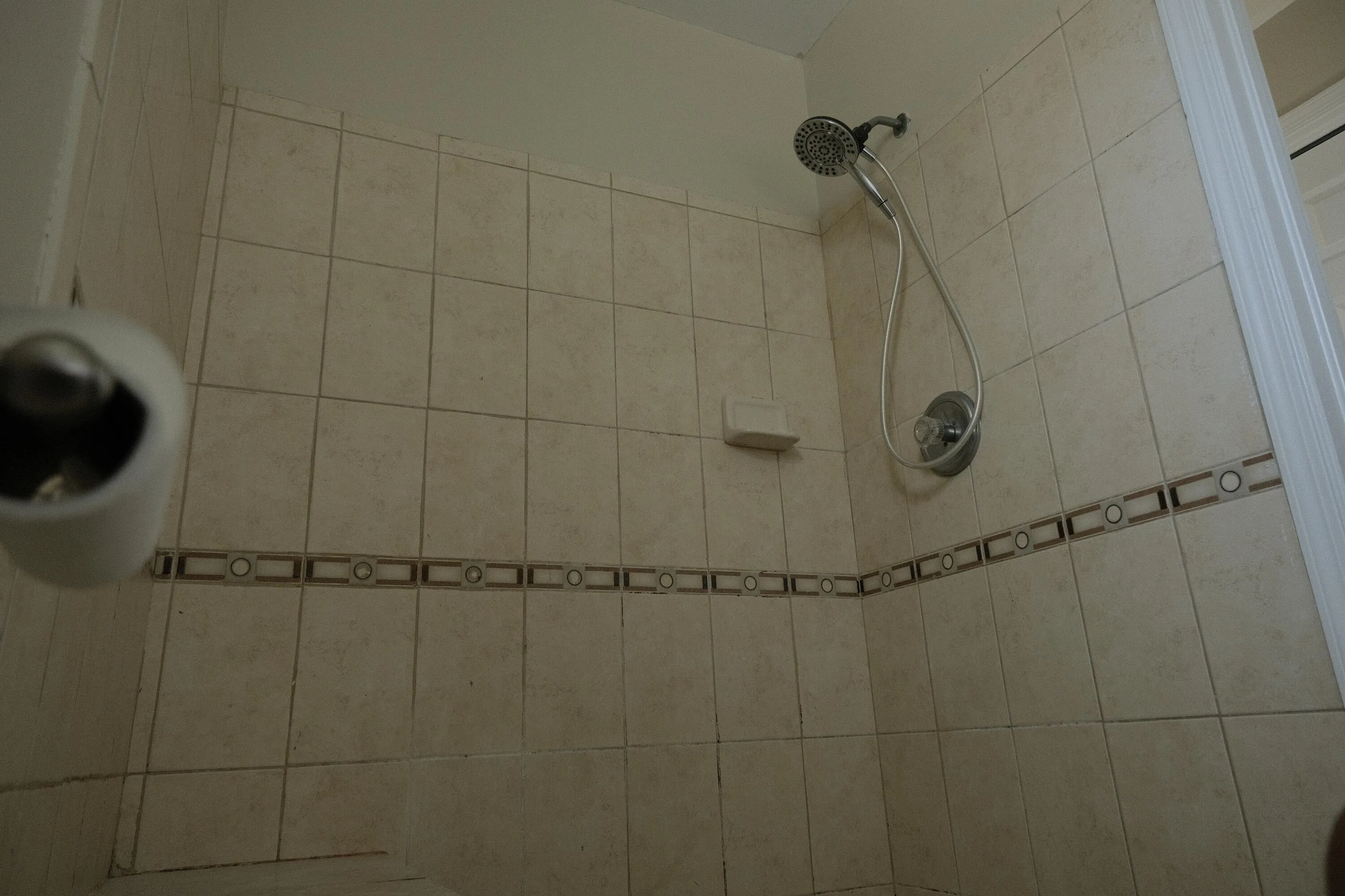 Shower stall featuring light-toned square tile, decorative accent tile band, and a hand-held showerhead with a diverter