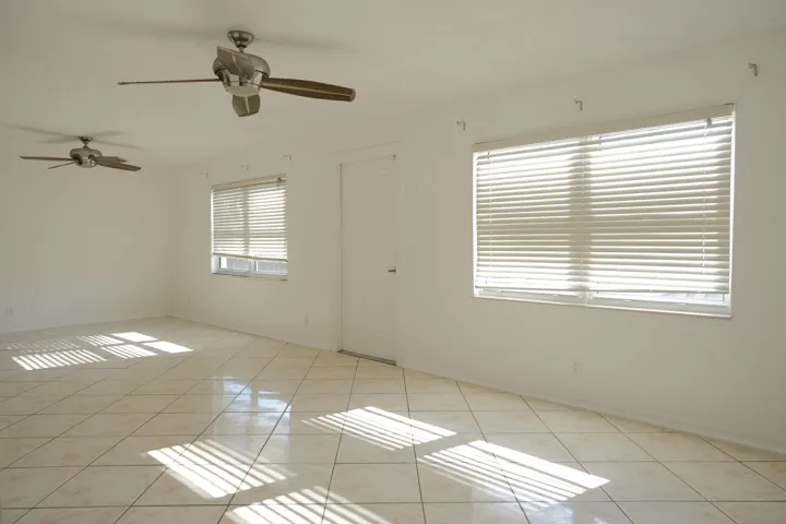 Spacious interior with tile flooring throughout, featuring two ceiling fans with integrated lighting, multiple windows with horizontal blinds, and a white interior door