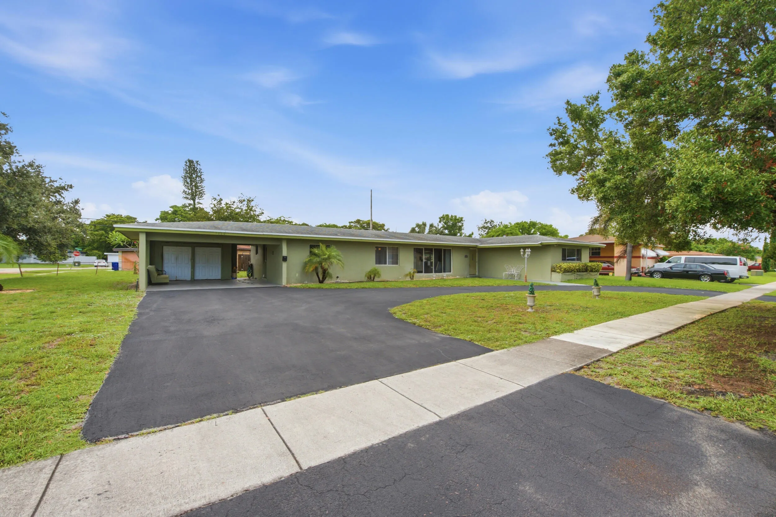 Single-story residence featuring a pale green exterior, expansive paved driveway, covered carport, and a concrete sidewalk