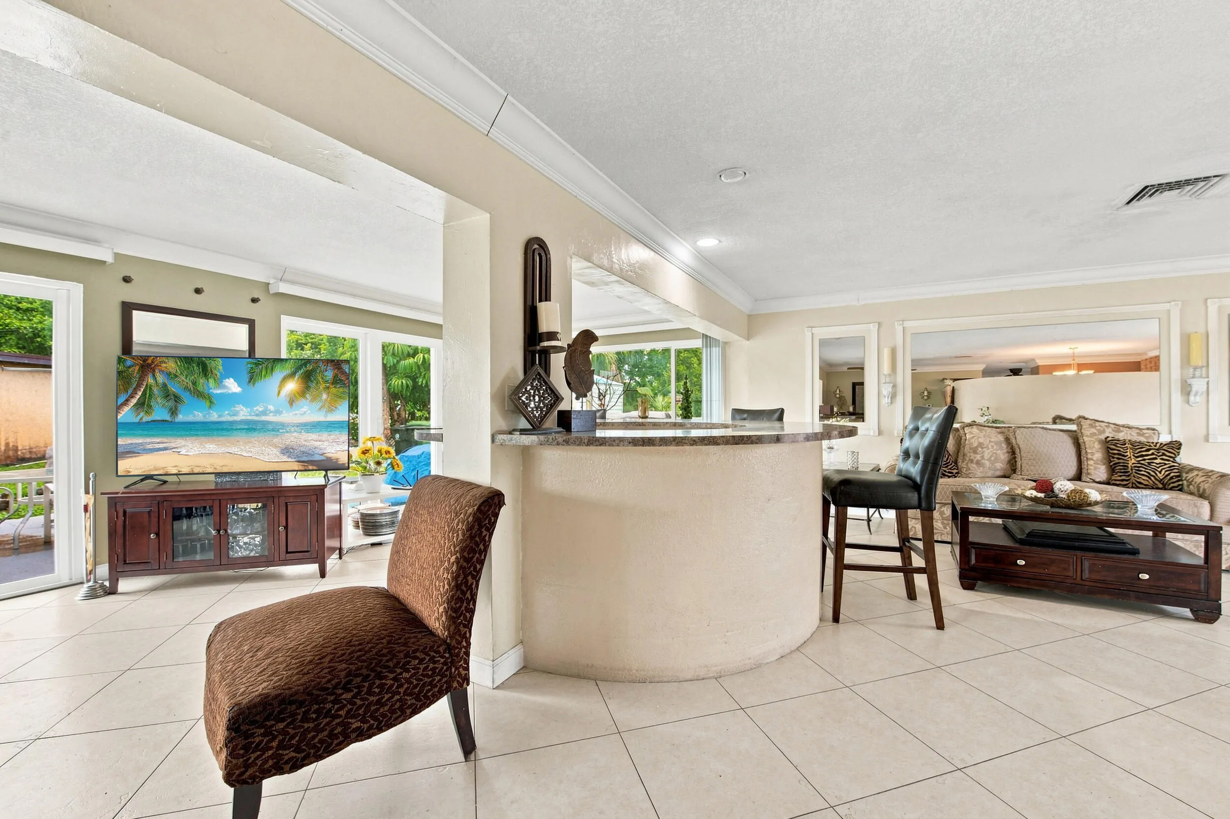Open-concept living space featuring light-toned tile flooring, a curved bar with a stone countertop, crown molding, recessed lighting, and expansive windows