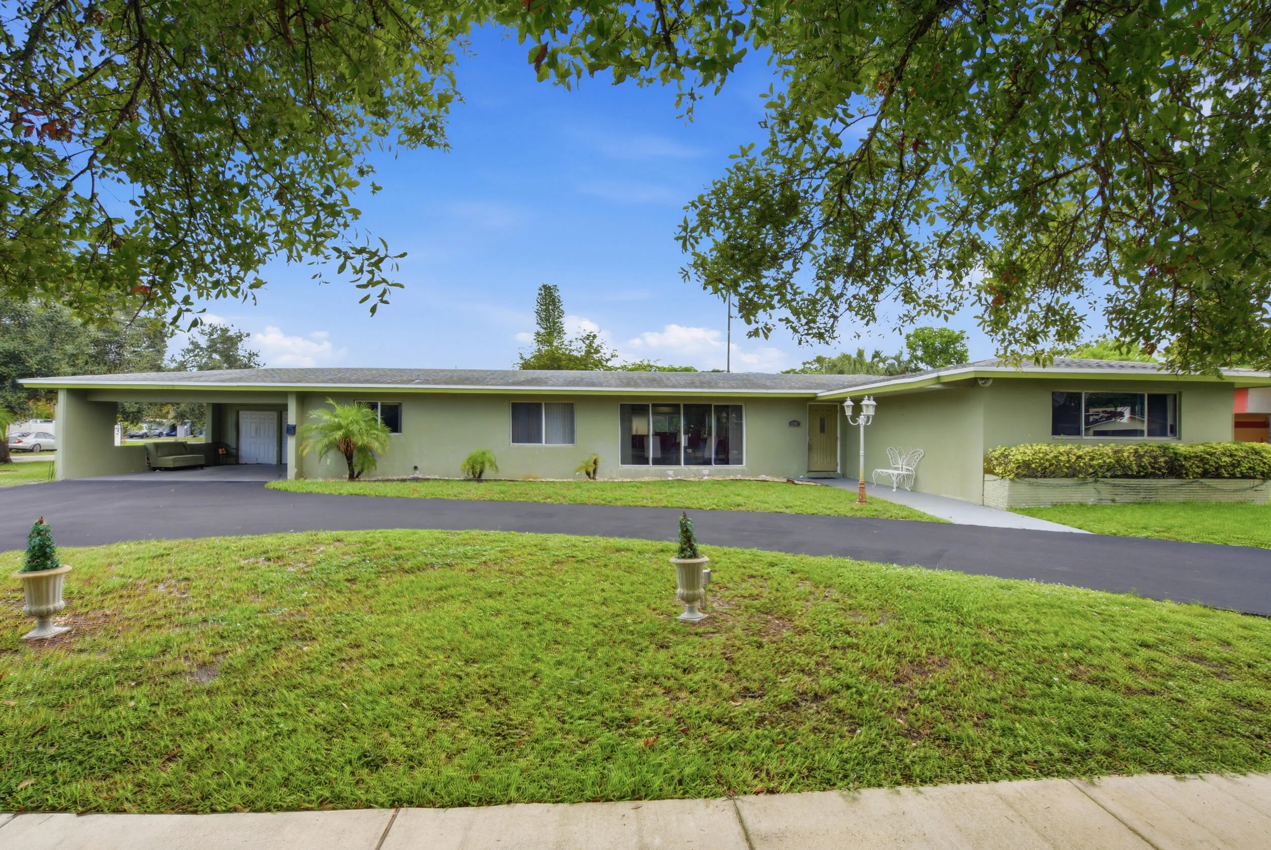 Light green exterior featuring a circular driveway, mature landscaping, and a covered carport