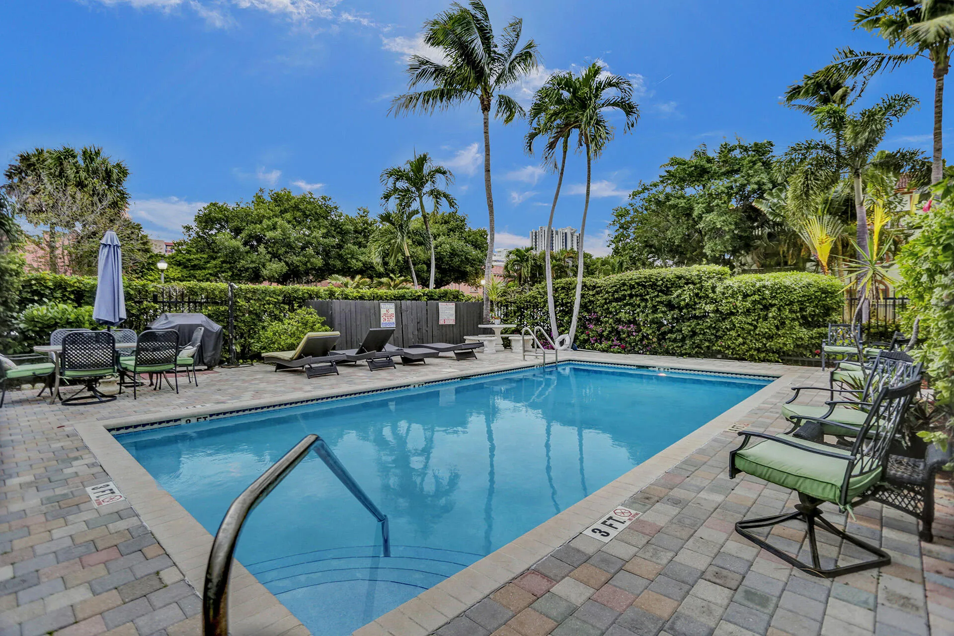 Outdoor swimming pool surrounded by paver decking, lush tropical landscaping, and mature palm trees
