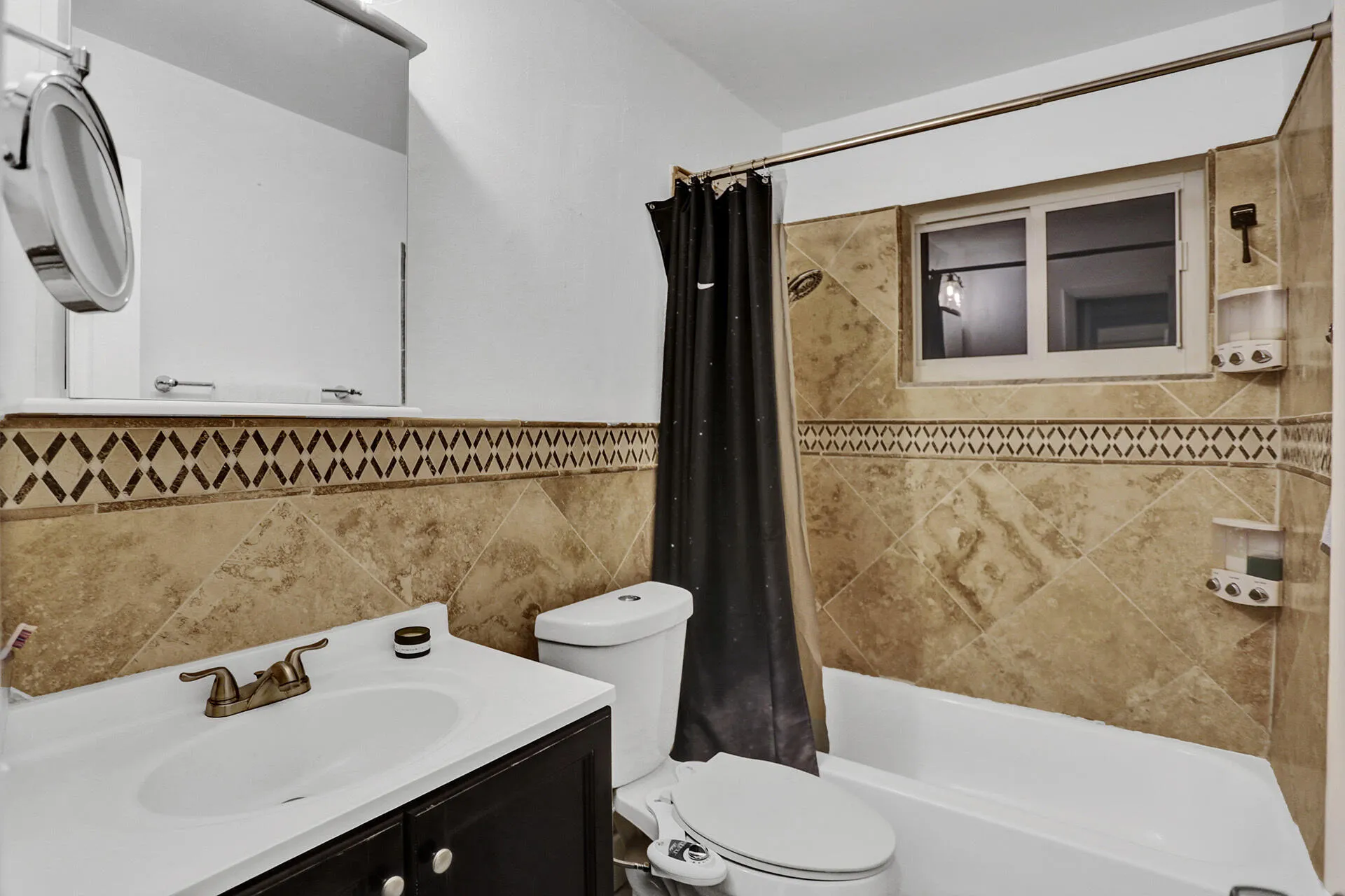 Bathroom featuring a tiled shower surround with a decorative accent band and an integrated window, a white bathtub, a toilet, a vanity with a white countertop and dark base, and a wall-mounted mirrored medicine cabinet