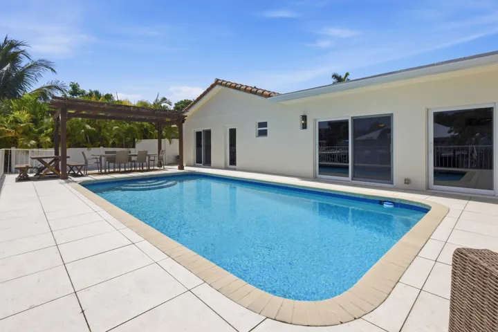 Expansive outdoor living area featuring a rectangular swimming pool with integrated steps, a large tiled patio, a wood pergola, and sliding glass doors providing access to the interior