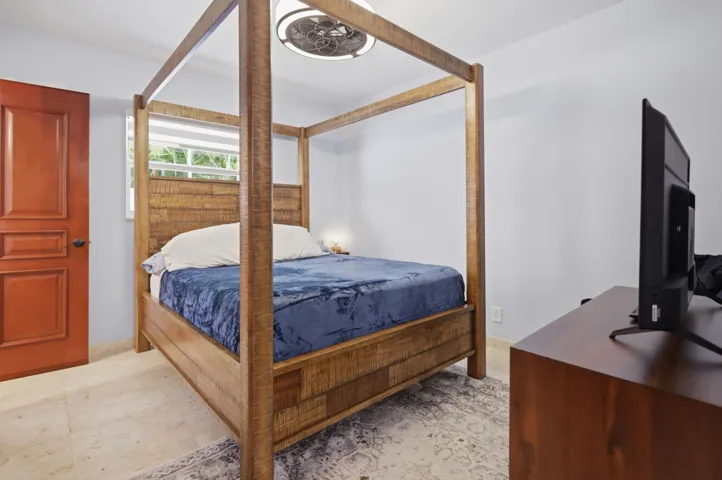 Bedroom featuring light gray walls, a window with outdoor foliage visible, a wood-finish four-poster bed frame, and cream-toned tile flooring