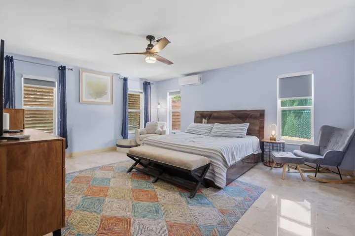 Bright interior space featuring light-toned tile flooring, multiple windows with blinds, a ceiling fan with integrated lighting, a contemporary wood-finish headboard, and light blue wall tones