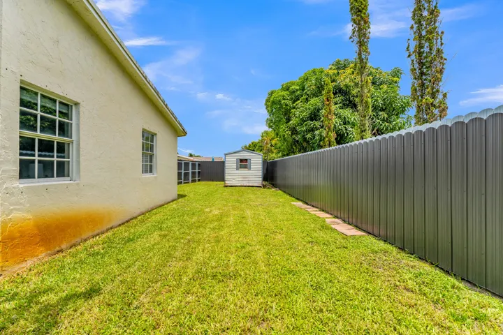 Expansive lawn area featuring a stucco-finish exterior, white-framed windows, a corrugated metal fence, and a storage shed