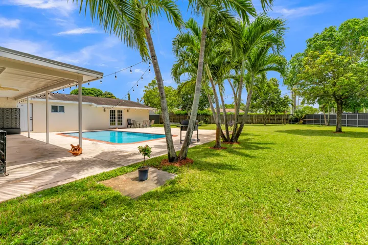 Expansive backyard featuring a rectangular swimming pool, a covered patio with string lighting, mature palm trees, a concrete patio, and a vast grassy area