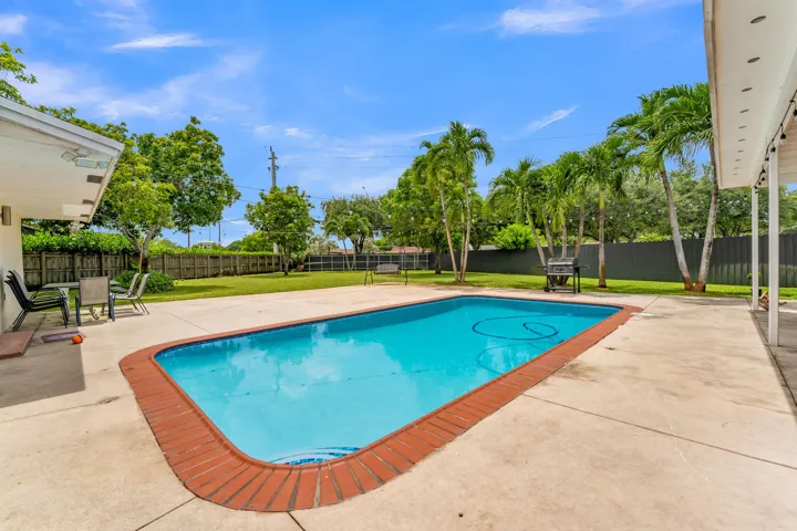 Expansive backyard featuring a rectangular swimming pool with brick coping, extensive concrete decking, a covered patio area, and mature palm trees