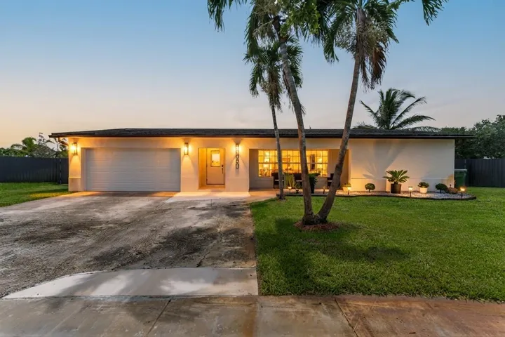 Single-story stucco residence with an attached garage, featuring exterior wall sconces, multiple palm trees, and a front lawn