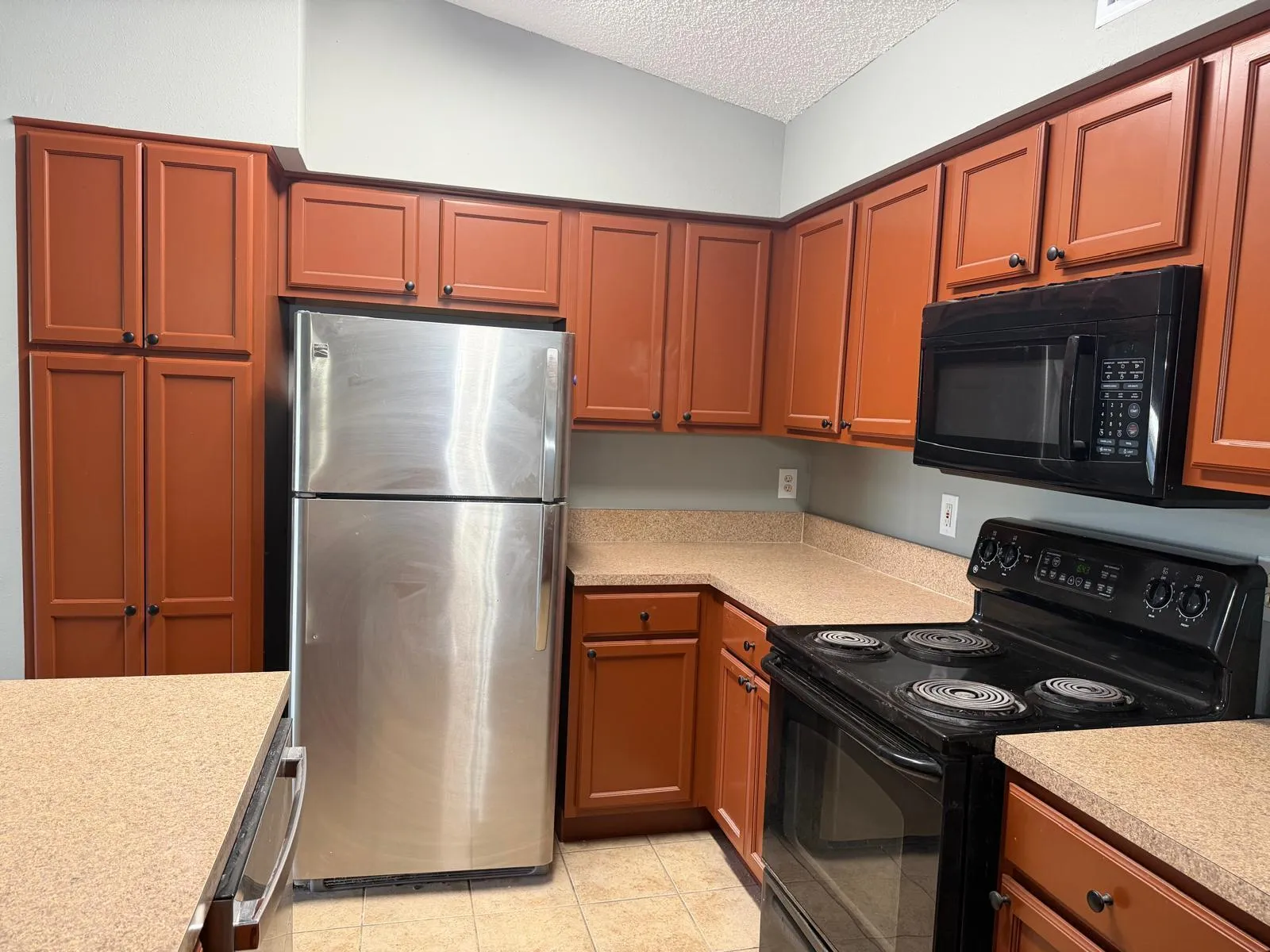 Kitchen featuring wood-finish cabinetry, light-toned countertops, and a full suite of appliances including a stainless steel refrigerator, black microwave, and black electric range