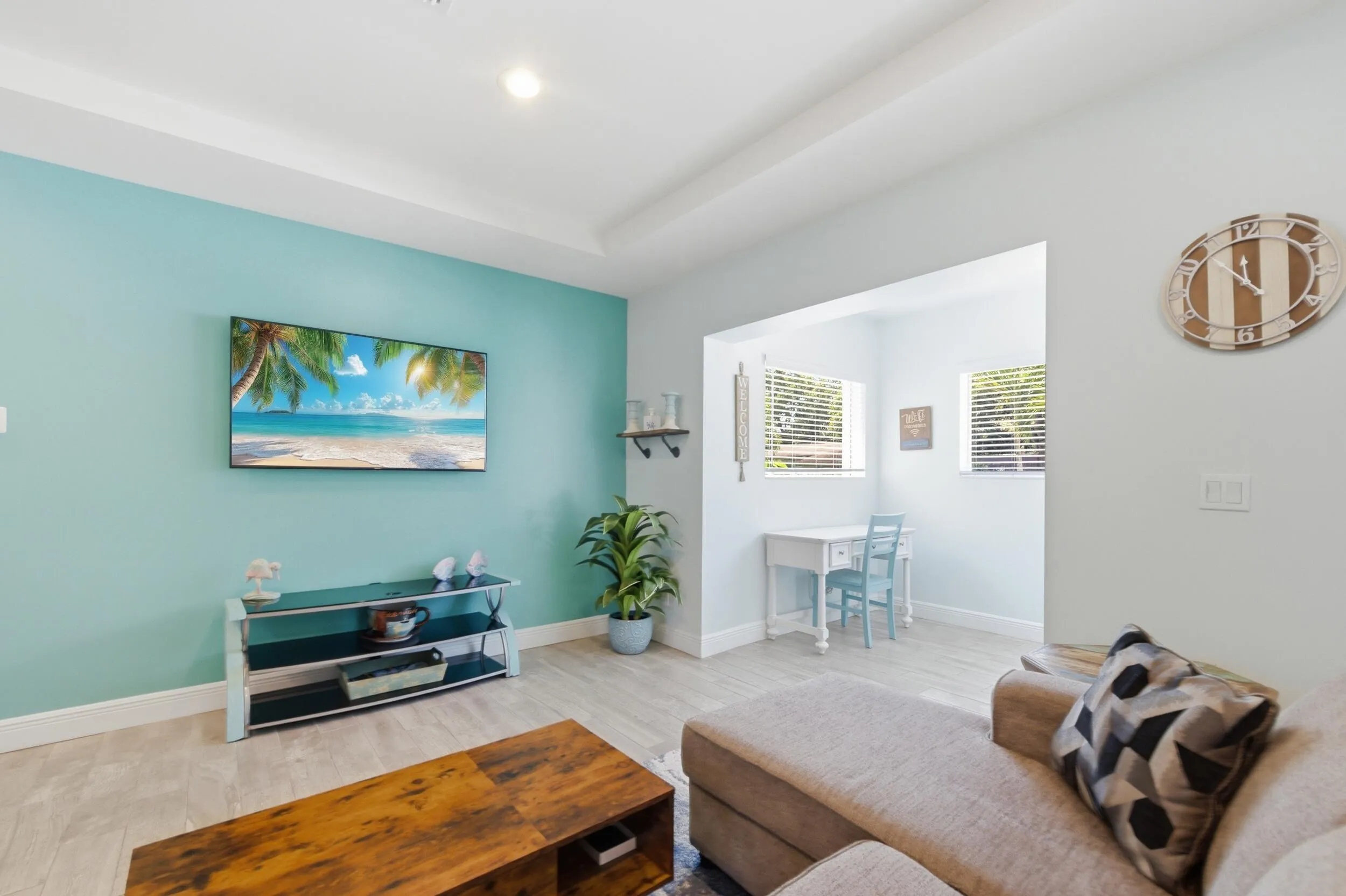 Living area featuring light-tone wood-finish flooring, a tray ceiling, and an open entryway to an adjacent space with two windows and horizontal blinds