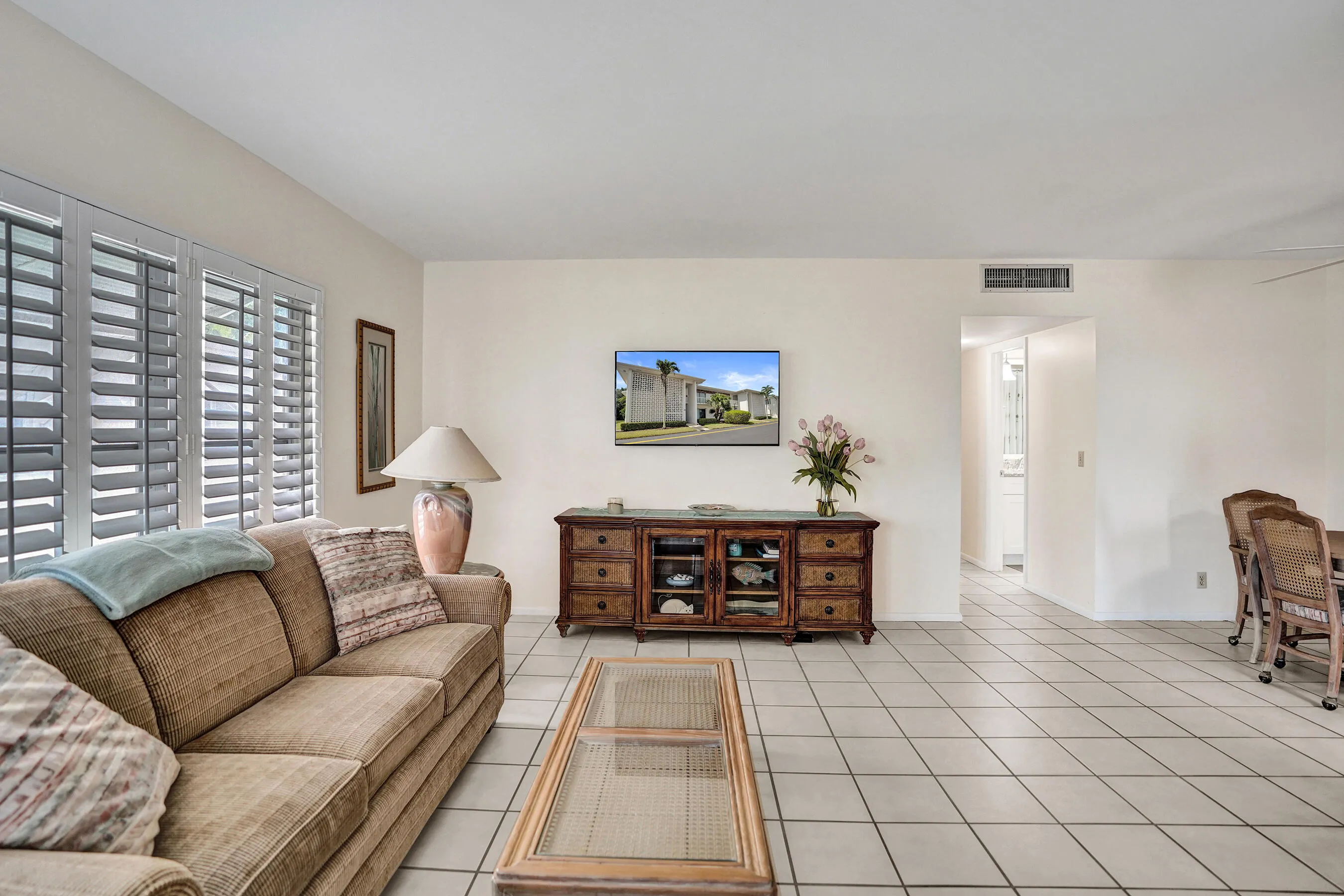 Large living area featuring white tile flooring, plantation shutters, and a wall-mounted display