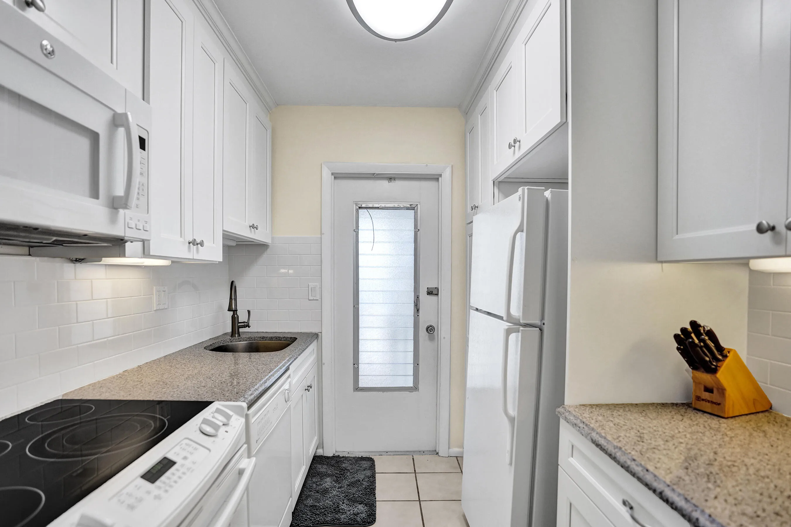 Galley kitchen featuring white cabinetry, speckled countertops, a subway tile backsplash, white appliances, and a rear exterior door with integrated blinds