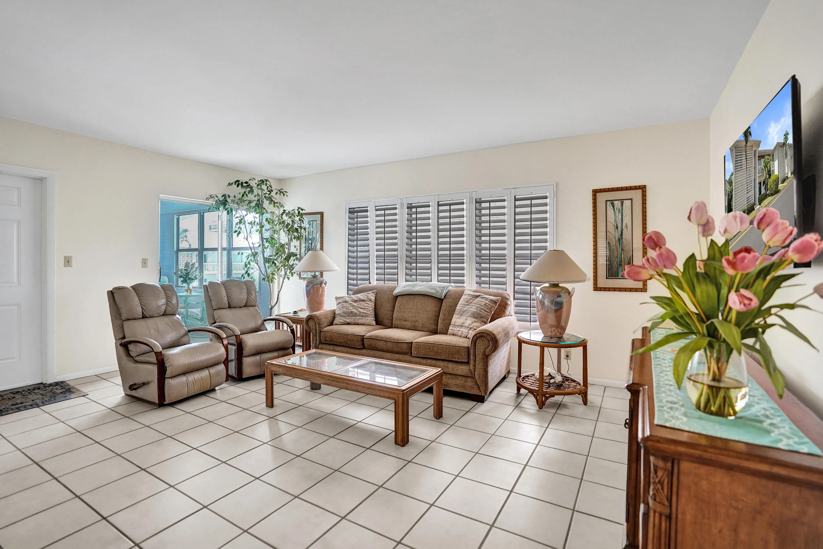 Spacious common area featuring light-toned tile flooring, white walls, and a prominent window with louvered shutters