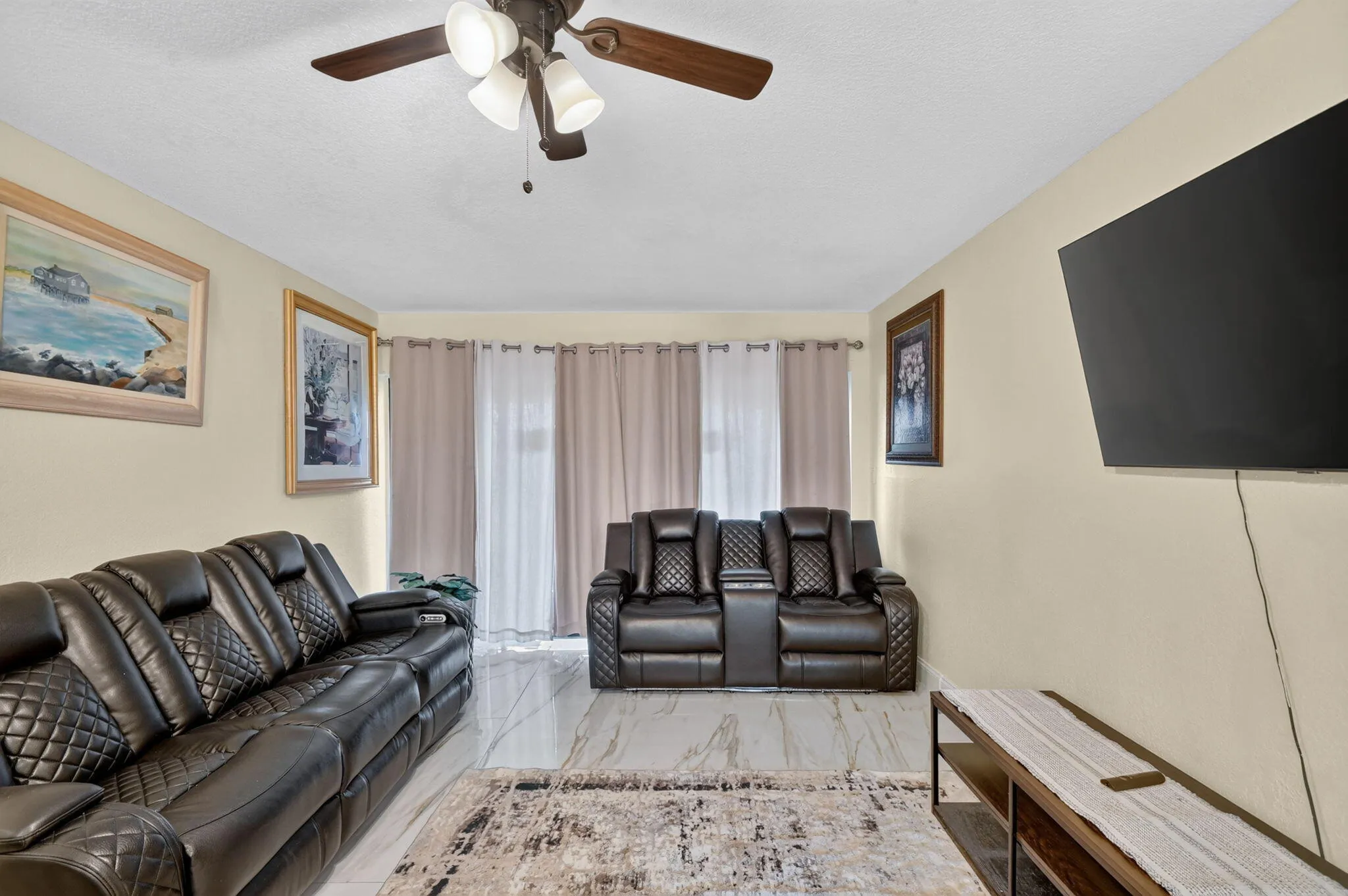 Living area featuring light-toned walls, a ceiling fan with integrated lighting, and neutral-toned curtains covering a light source