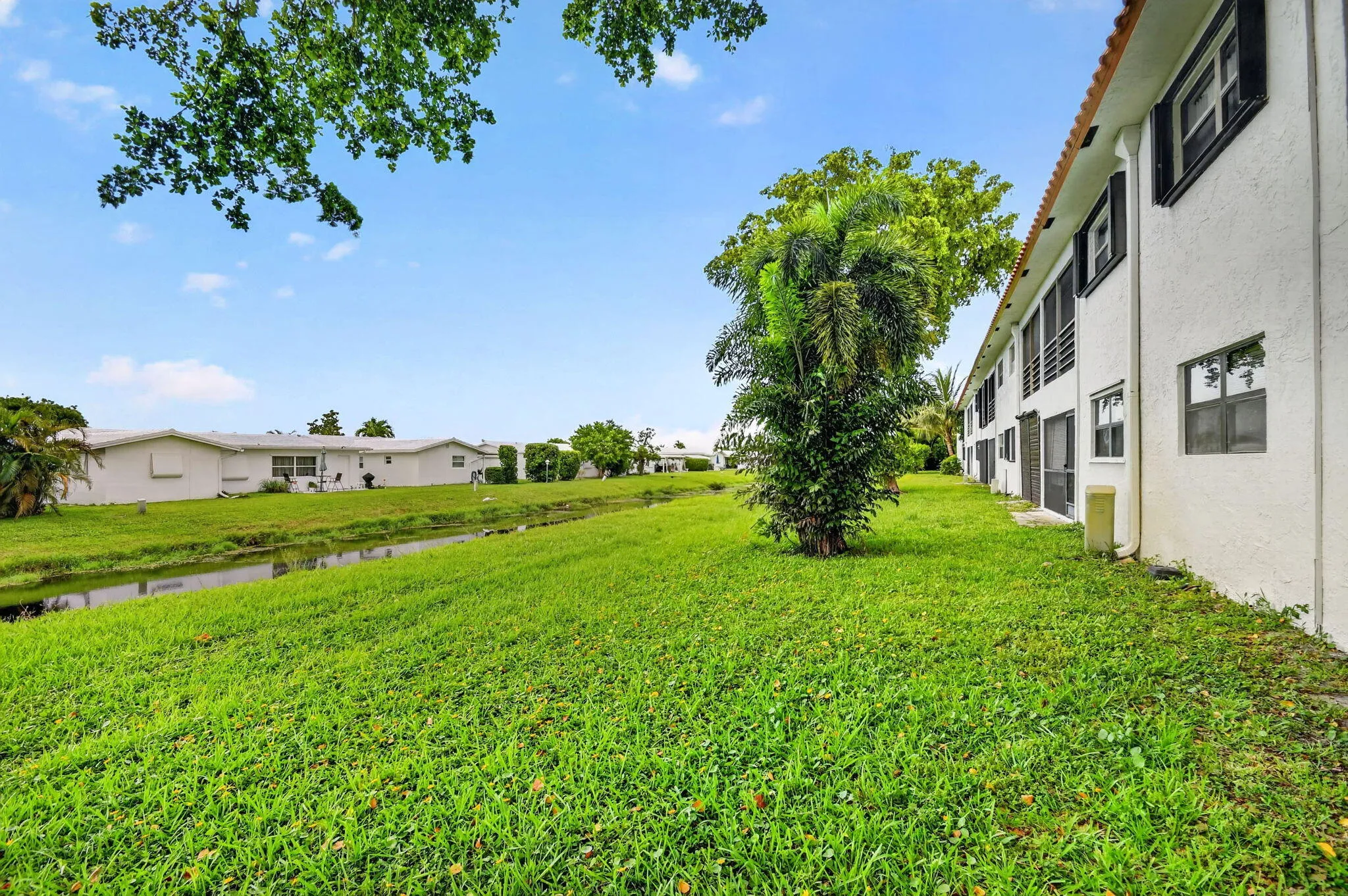 Expansive green lawn bordering a water feature, with a multi-story building featuring white stucco siding, dark shutters, and a red tile roof