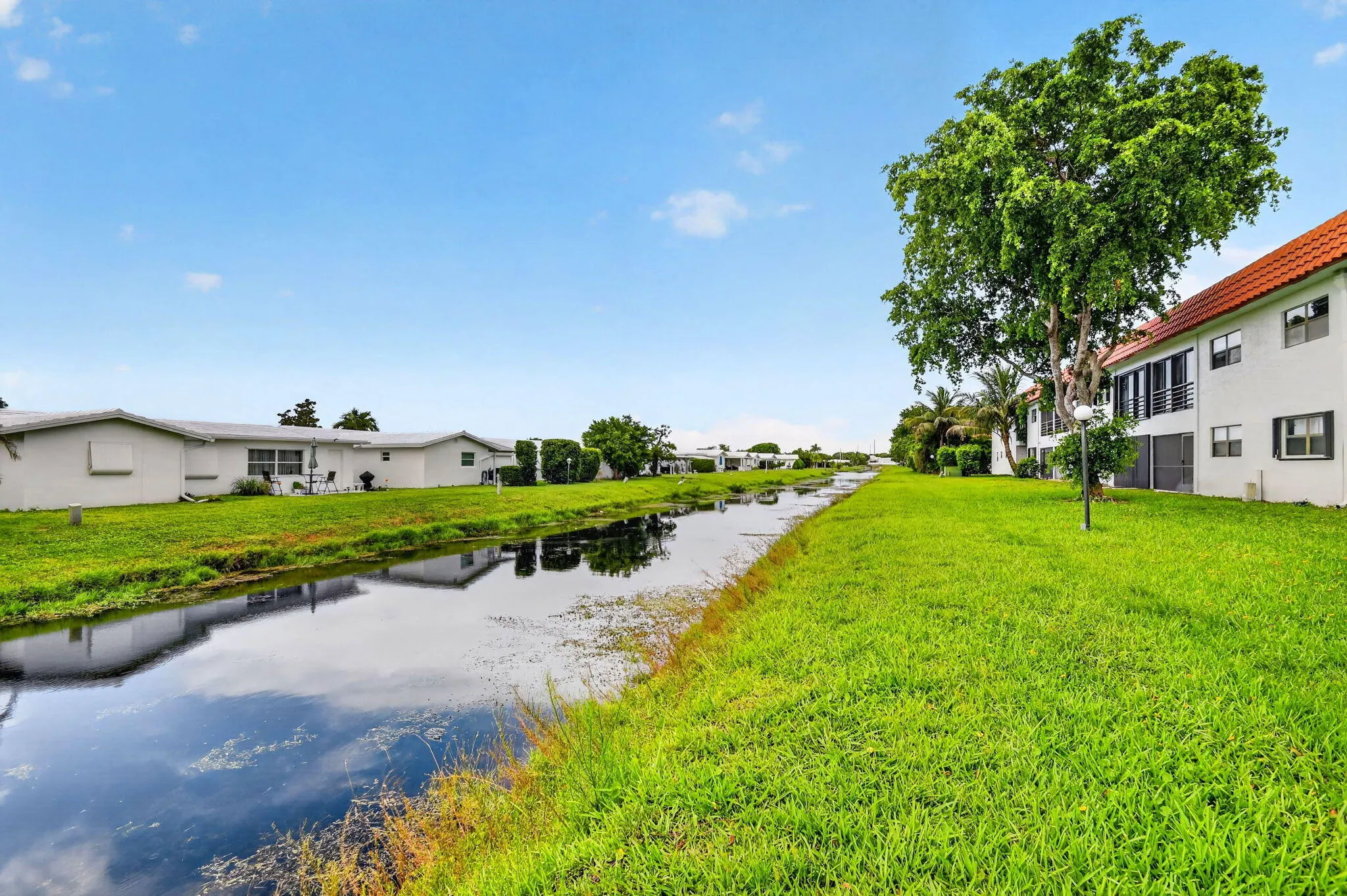 Waterfront property featuring a canal, expansive green lawn, mature trees, white exterior facades, and red tile roofs