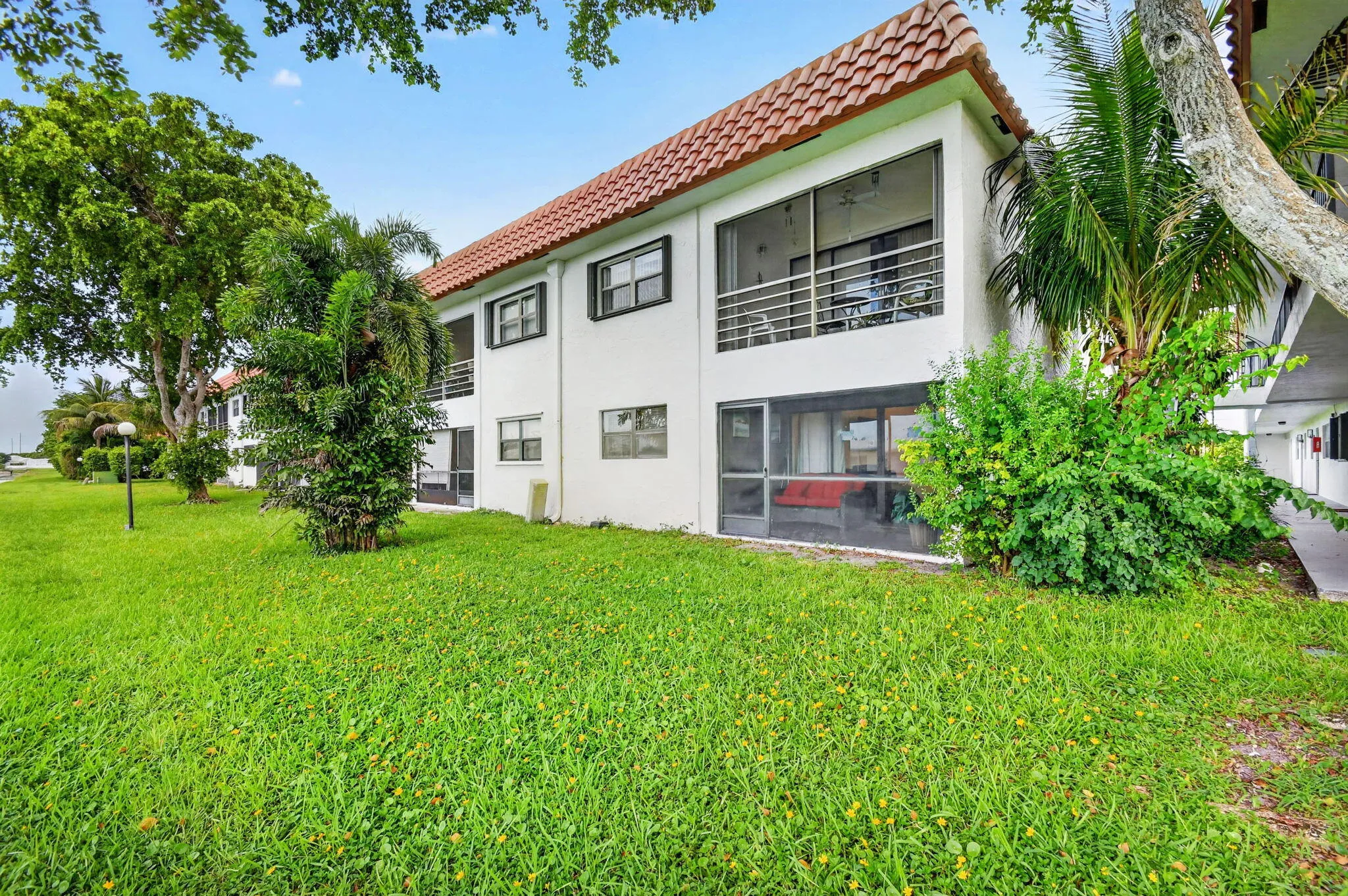 White stucco exterior with a terra cotta tile roof, featuring screened upper and lower balconies, black-framed windows with exterior shutters, and surrounding mature landscaping
