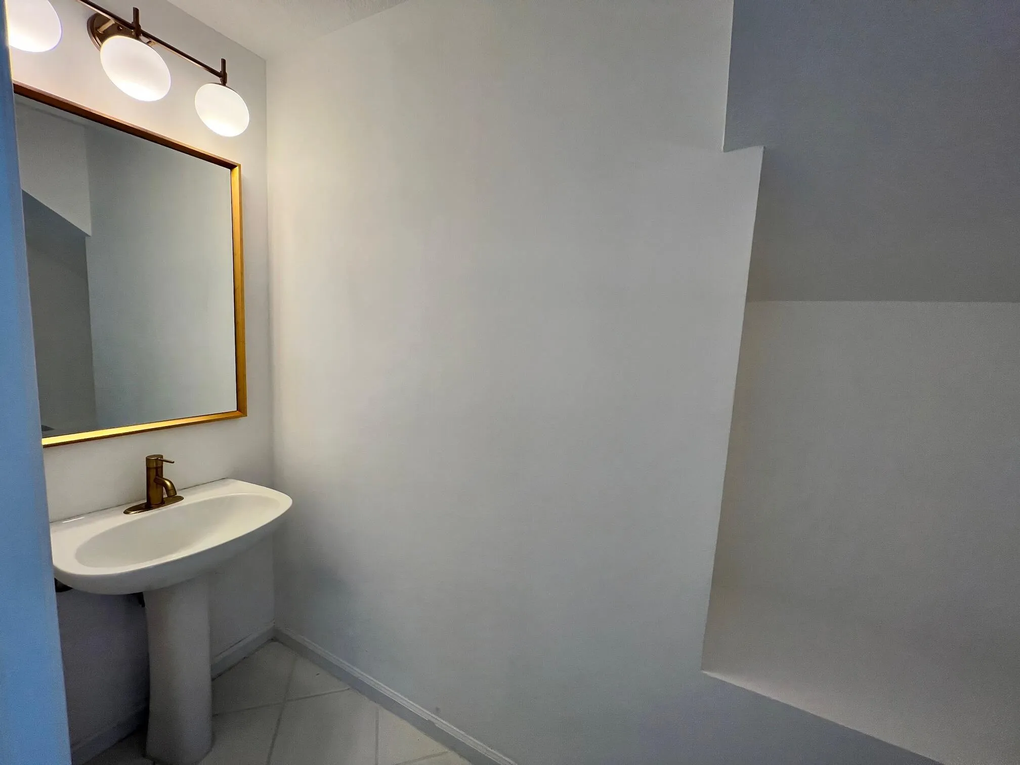 Pedestal sink with a brass-finish faucet, framed mirror, and a three-bulb vanity light with frosted globes